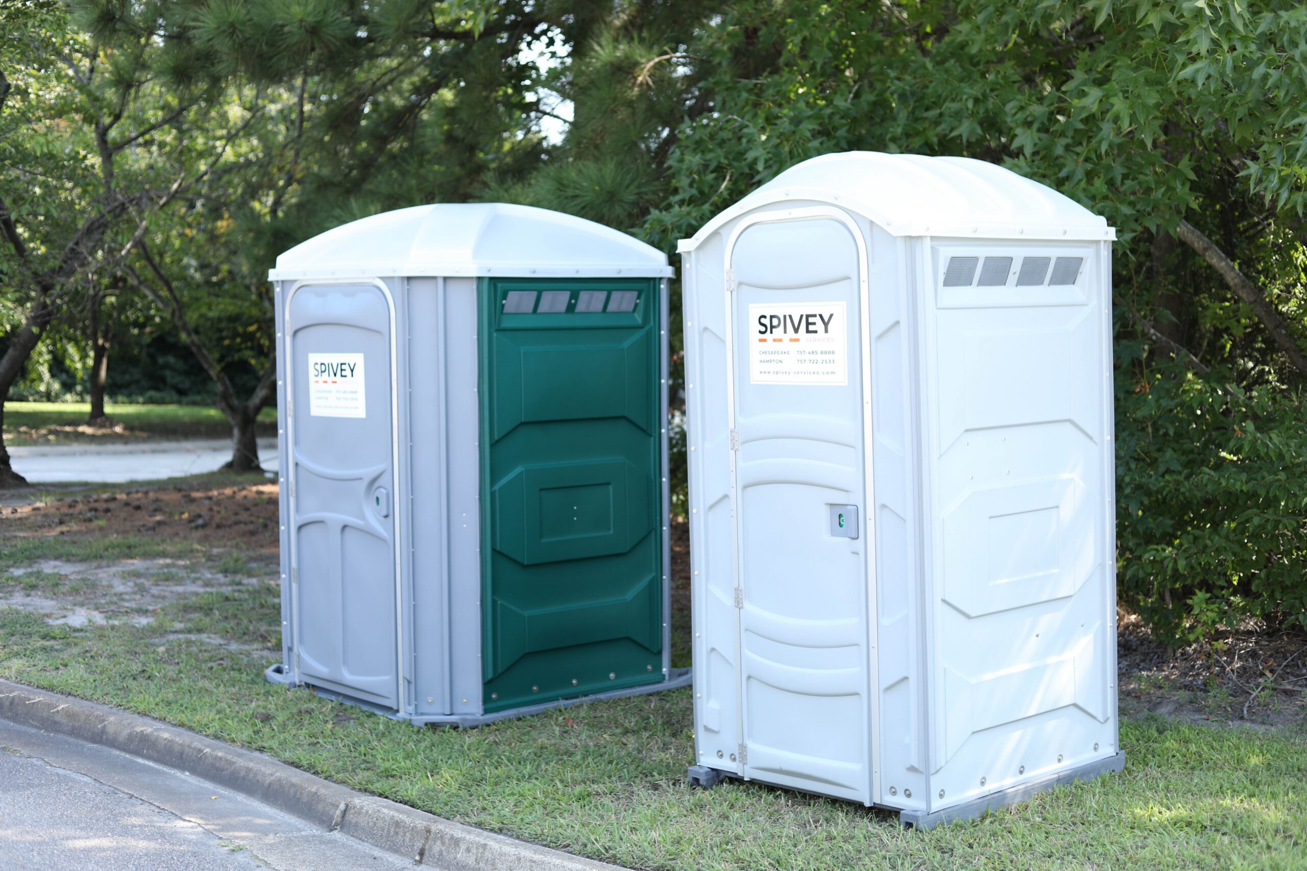 Spivey porta potty rental of a handicap-accessible unit and a standard unit sitting on grass between a curb and some trees